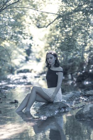 Dreamy nature photo of someone sitting on rocks by a reflective stream in soft lighting.