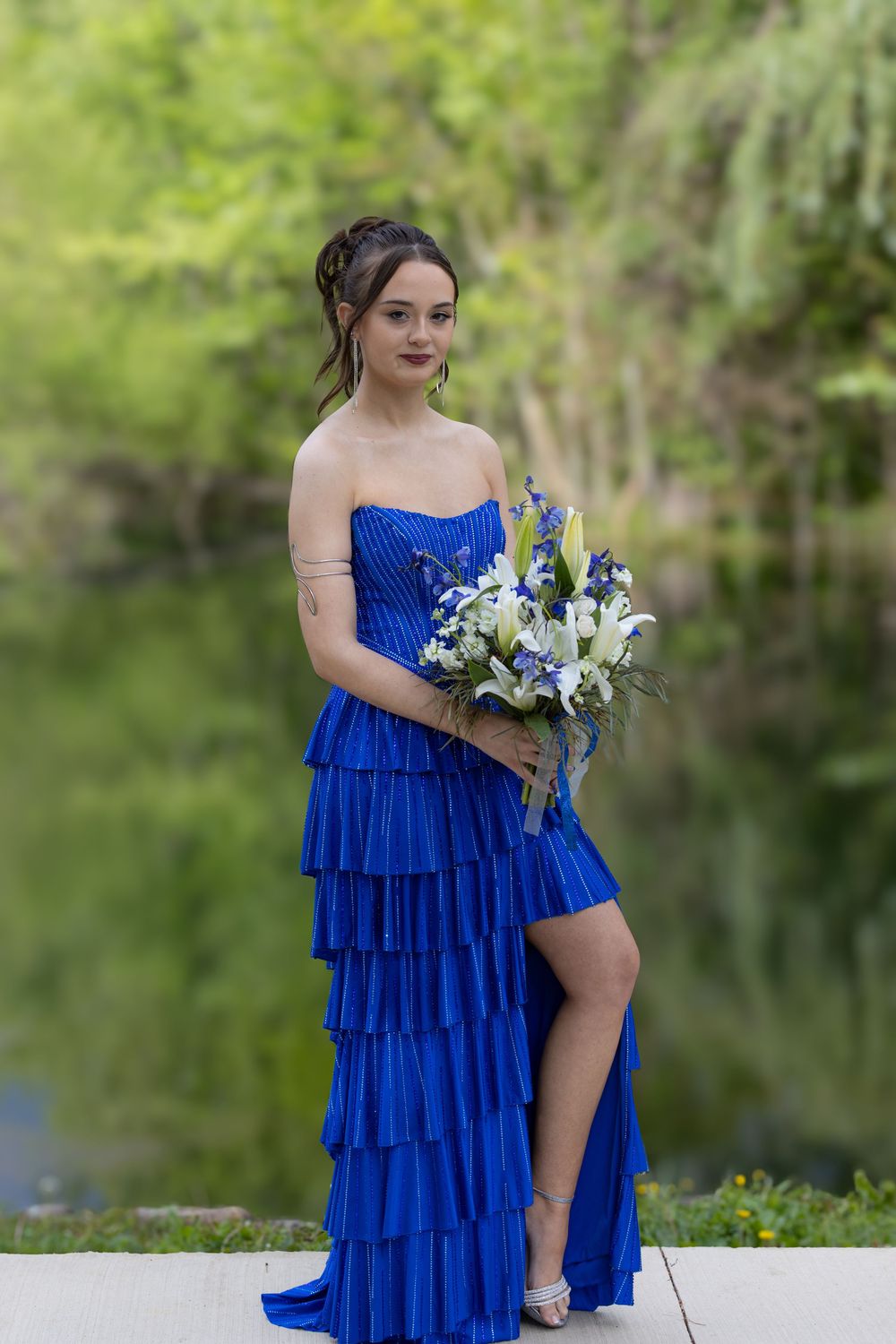 Person in a vibrant royal blue tiered strapless gown poses with white floral bouquet by a tranquil lake.