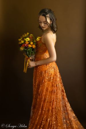 A dramatic side view portrait of someone in a flowing orange sequined gown with flowers.