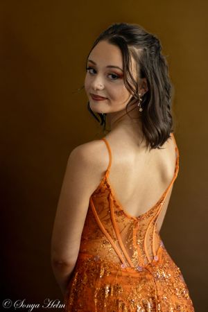 Elegant portrait of woman in sparkling orange formal dress looking over her shoulder against brown background.