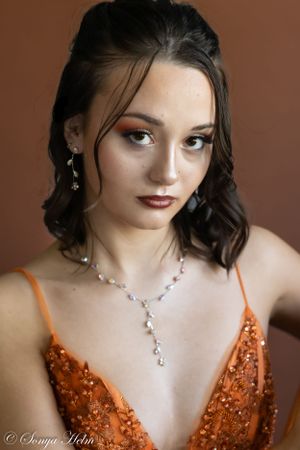 Glamorous portrait of model in orange sequin gown wearing delicate crystal necklace against brown backdrop.