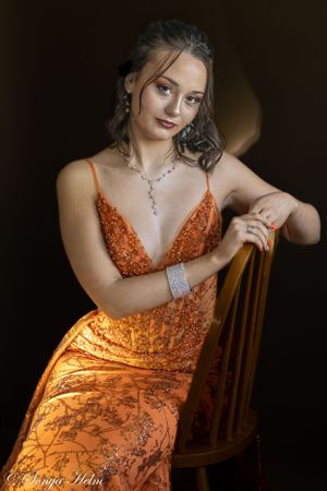 Dramatic portrait of model in orange sequin gown posing elegantly on wooden chair.