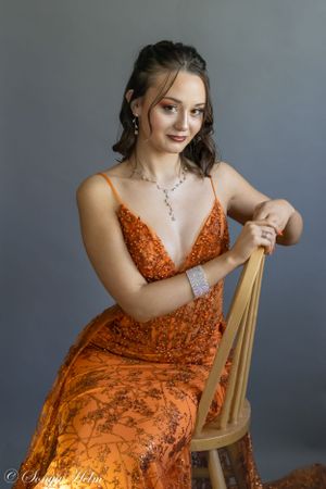Sophisticated portrait showing model in orange formal gown posed on vintage chair.