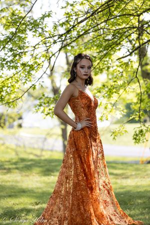 Outdoor fashion portrait under green trees featuring flowing orange sequin gown.