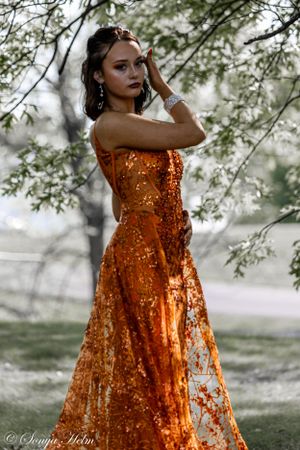 Stunning sequined orange evening gown photographed outdoors with soft natural lighting and tree branches in background.