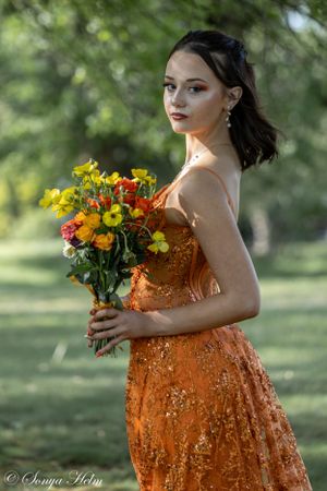 Model in sparkling orange evening gown poses with vibrant yellow and orange flower bouquet in outdoor garden setting.