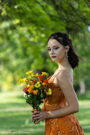 Woman wearing sequined orange dress holds colorful summer bouquet against lush green garden backdrop.