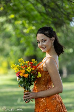 Graceful figure in shimmering orange dress smiles while holding yellow and orange wildflower bouquet.
