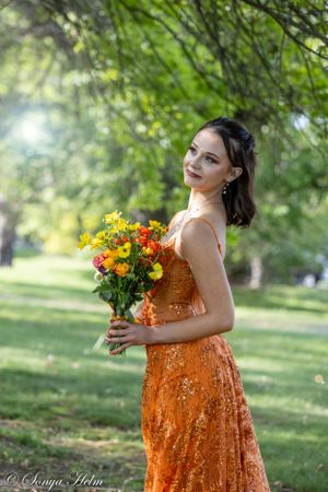 Elegant woman in orange sequin gown holds bright floral arrangement while posing in sunlit park.