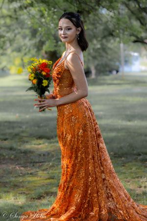 Full-length portrait of woman in flowing orange sequined gown holding bouquet in park setting.