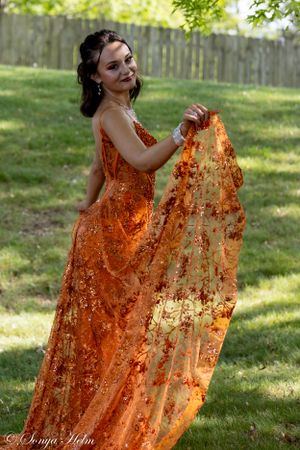 Woman in orange sequined gown twirls dress fabric in outdoor garden setting.