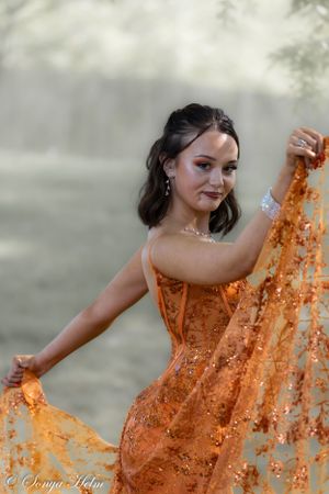 Woman in orange sequined gown playfully lifts dress fabric in misty garden setting.