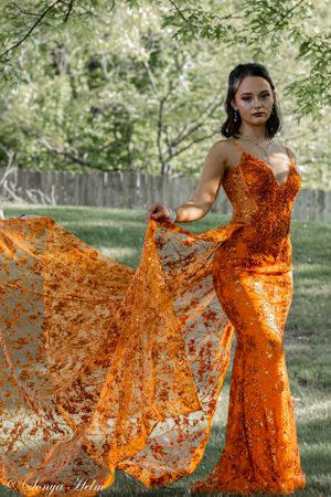 A fitted orange sequin gown with flowing fabric panel photographed in natural lighting.