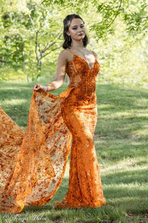 A form-fitting orange sequined gown showcased against green garden foliage.