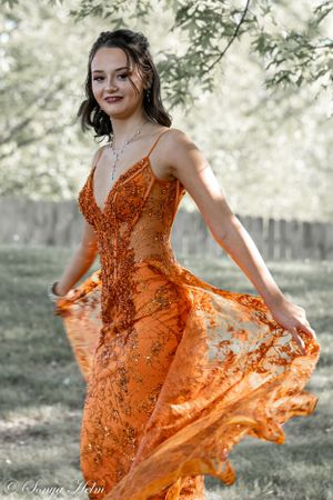 An orange sequined gown twirls gracefully in a sun-dappled garden setting.