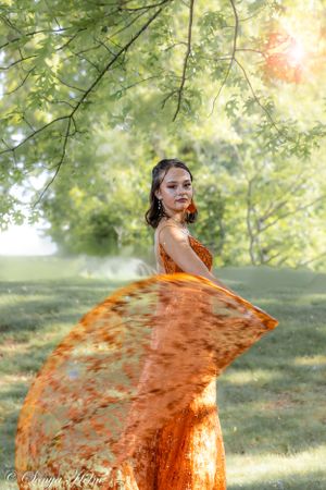A flowing orange evening gown creates movement against green summer foliage.