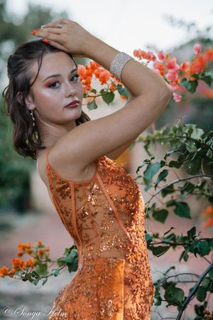 A woman in a sparkly orange gown poses elegantly among blooming coral flowers in a garden setting.
