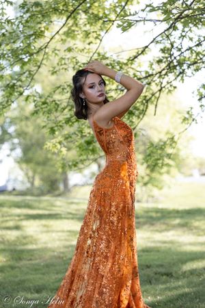 A flowing orange sequined gown creates drama against a sunlit natural garden backdrop.