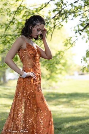 A pensive pose in a sparkling orange gown set against a soft, natural garden background.