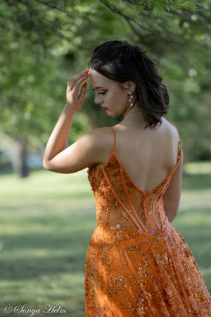 A model in an elegant orange sequined dress stands in profile against a blurred natural background.