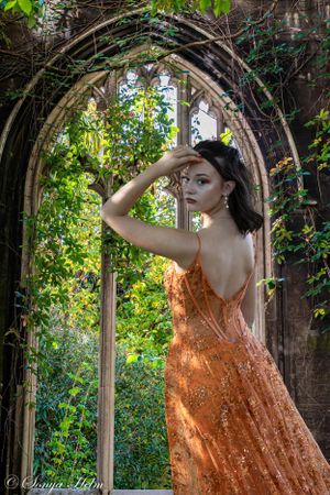 A stunning orange formal dress is photographed through a gothic archway covered in climbing vines.