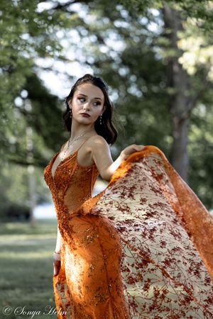 An elegant orange gown with intricate detailing is captured in motion against trees.