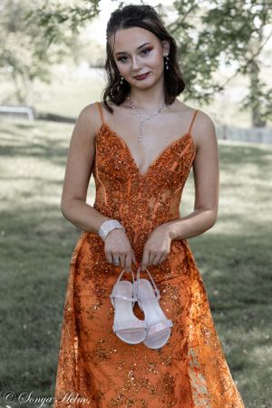 Woman in sparkly orange formal dress holds white shoes while posing outdoors in a garden setting.