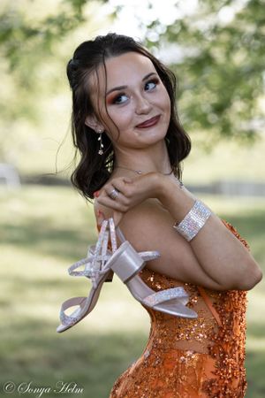 Woman in glittering orange dress holds shoes while striking a playful pose against a blurred nature backdrop.