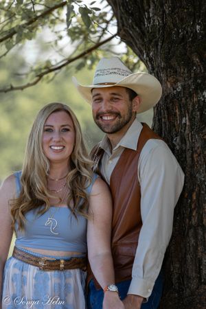 A western-styled couple poses together by a tree trunk in casual country attire.