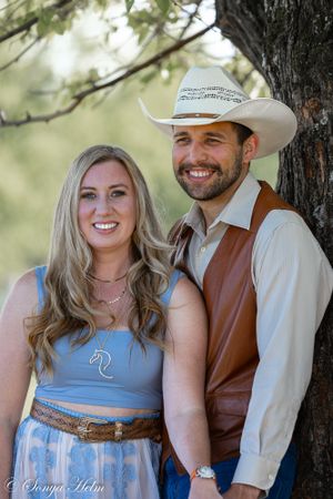 A couple in western wear smile while standing against a large tree trunk outdoors.