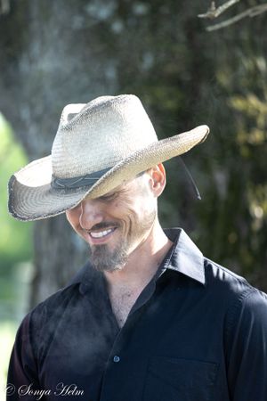 A cowboy in a black shirt and light-colored hat smiles outdoors on a sunny day.