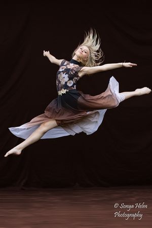Graceful dancer leaps through the air in flowing dress with floral bodice against dark background.