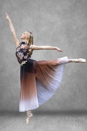 Ballet dancer performs elegant pose in ombre dress with floral top against textured grey wall.