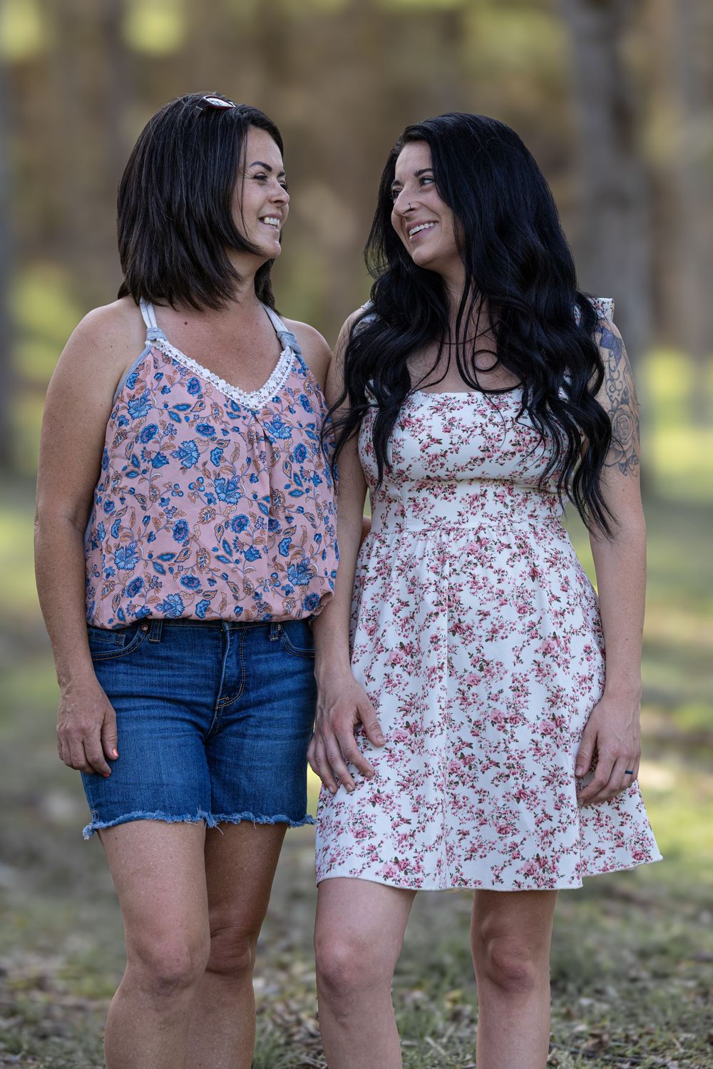 Two people in summer outfits stand close together and share joyful smiles while spending time in a sunny outdoor setting.
