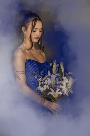 A person in a blue strapless dress holds an elegant bouquet of white lilies and flowers in a dreamy misty setting.