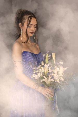A figure in a purple gown holds a white lily bouquet against a dreamy, misty gray background in an ethereal setting.