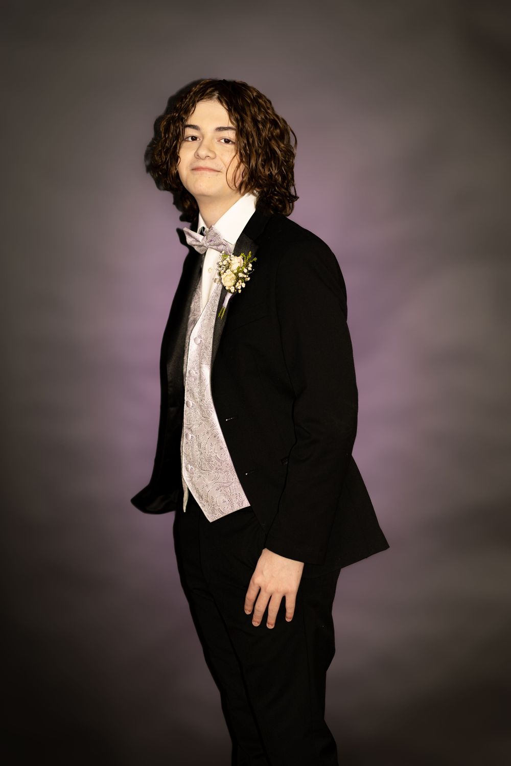 A young person in a formal black suit and silver vest poses for a portrait against a dark background with curly dark hair.