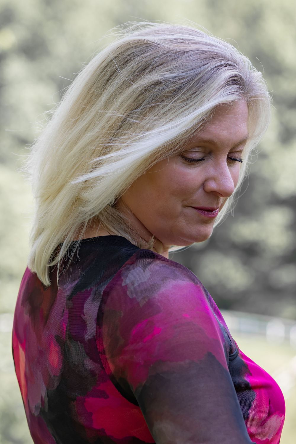 A person with blonde hair wearing a magenta dress poses against a soft-focus nature background.
