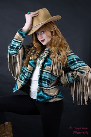 Model wearing turquoise and tan fringed western jacket with brown cowboy hat poses dramatically against dark background.