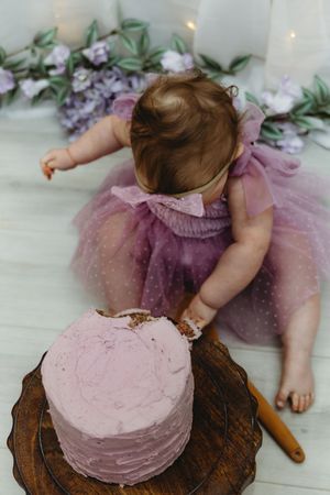 Baby girl in purple tutu dress reaching into pink birthday cake on wood stand.