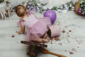 Toddler girl in pink tutu dress smashing first birthday cake with purple balloons.
