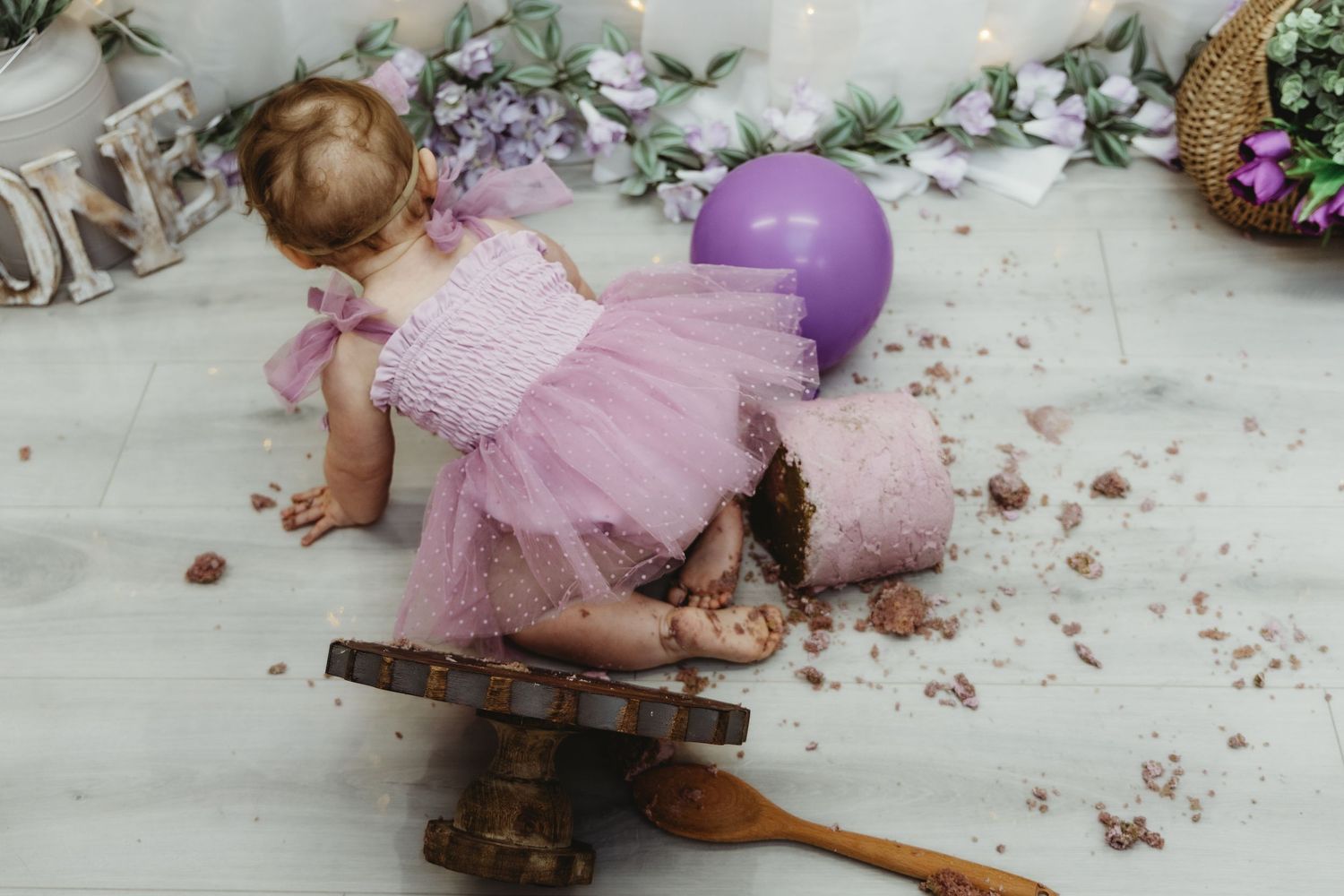 Toddler girl in pink tutu dress smashing first birthday cake with purple balloons.