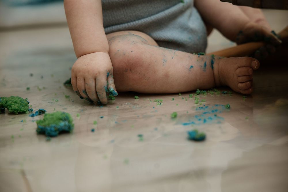 Baby hands and legs covered in blue and green cake smash frosting sitting on floor.