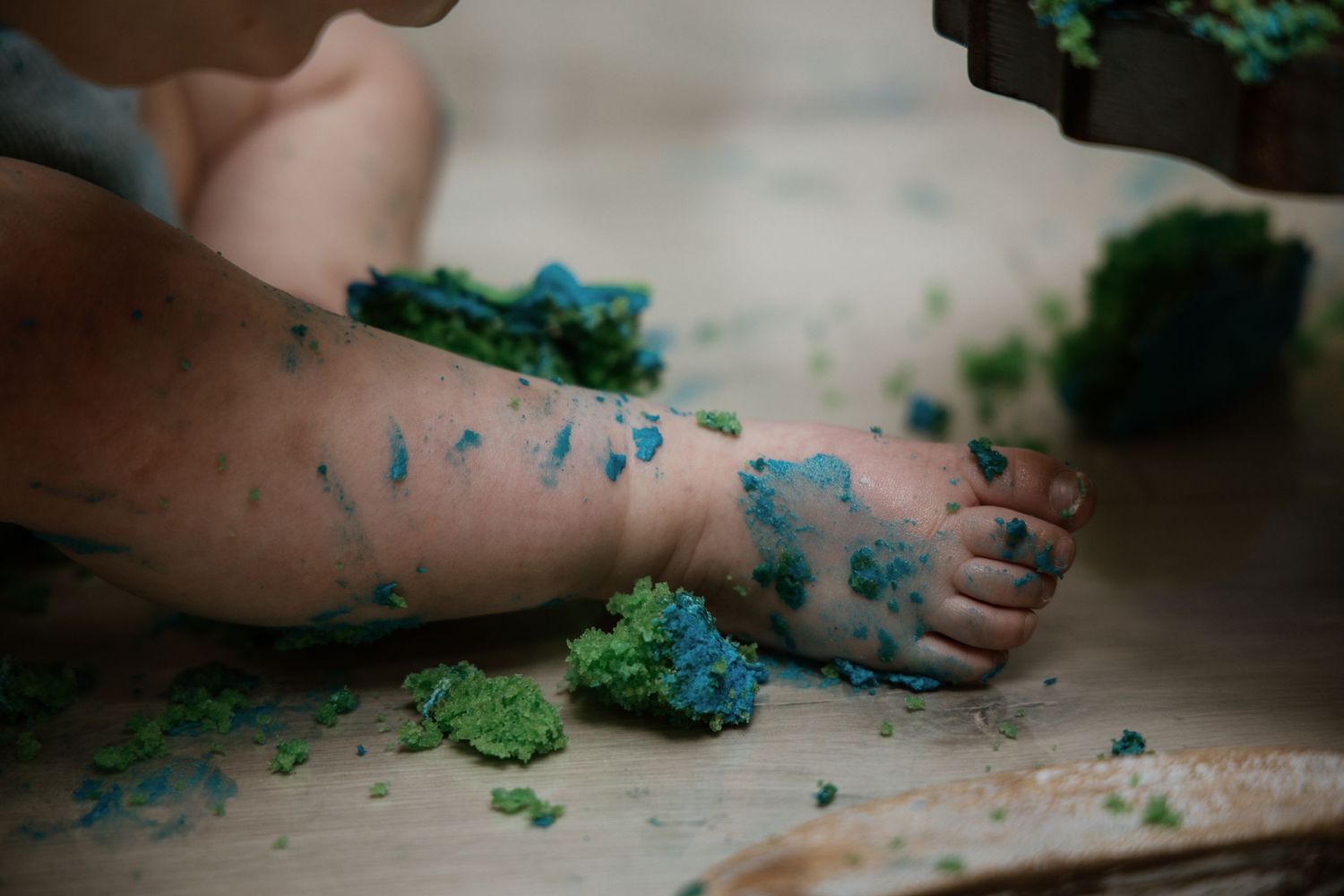 Close-up of baby feet and legs with colorful cake smash frosting on wooden surface.