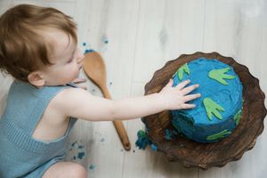 Overhead view of toddler in gray romper touching blue and green birthday cake.