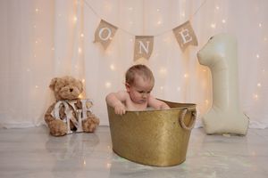 Baby sitting in a golden tub at a first birthday photoshoot with teddy bear and ONE banner.