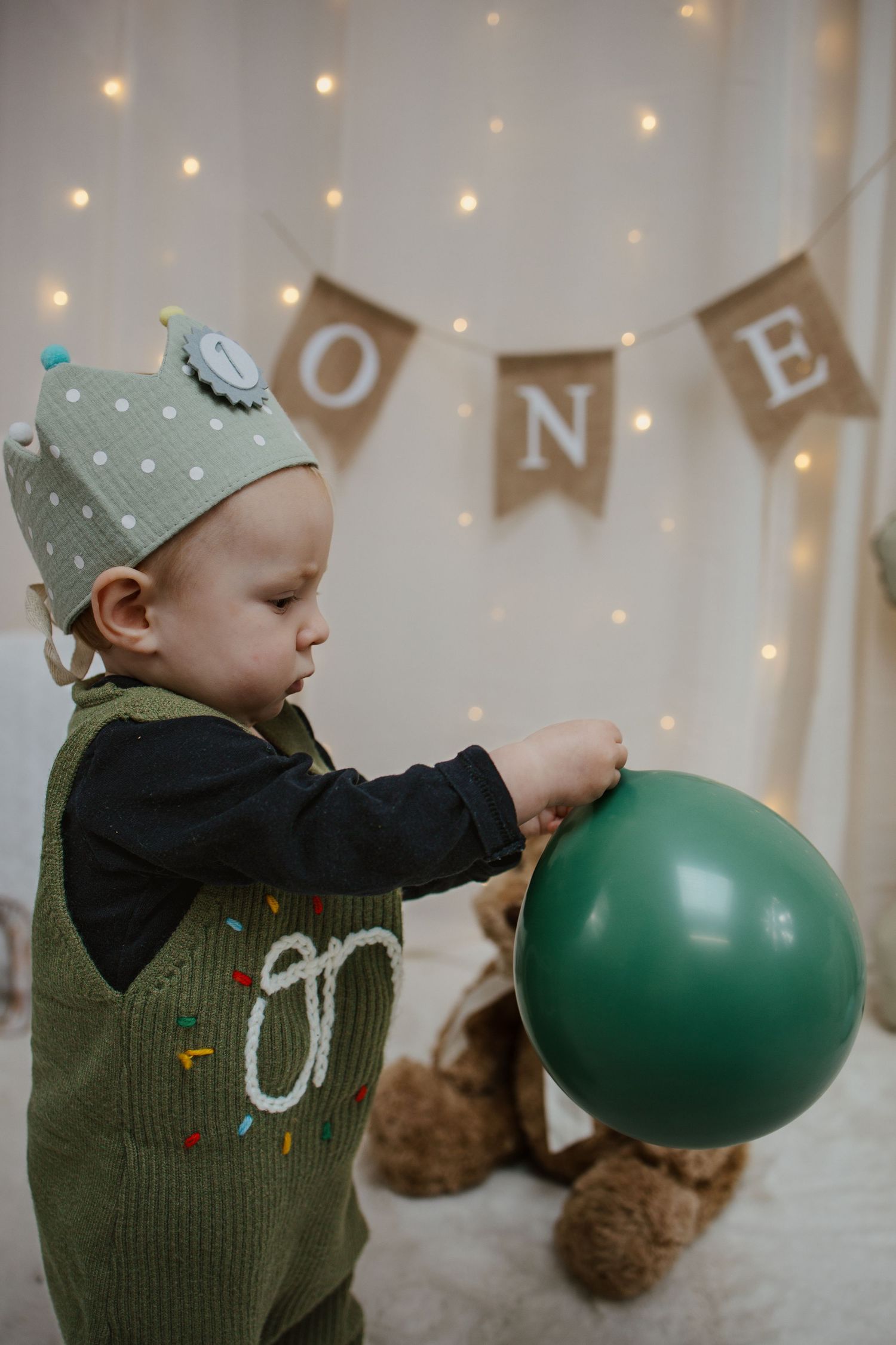Baby wearing a crown hat and green overalls plays with a balloon at a first birthday party.