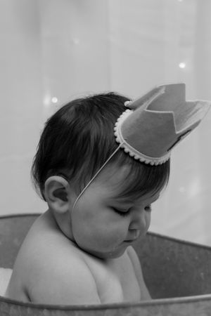 Black and white photo of a baby wearing a crown headband sitting in a metal tub.