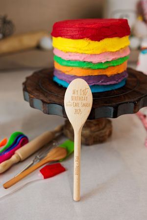 Rainbow layered smash cake on wooden stand with engraved My 1st Birthday wooden spoon.