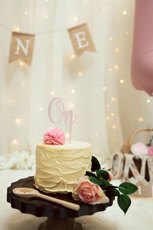 Elegant first birthday cake with pink flowers and One topper on a wooden stand with fairy lights.